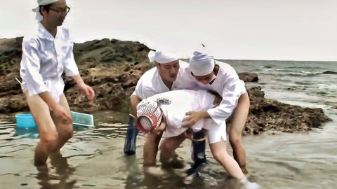 Japanese woman in white robe helped by two men in water on rocky beach