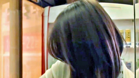 Japanese woman with long black hair in store uniform turning profile in front of orange-lit fridge