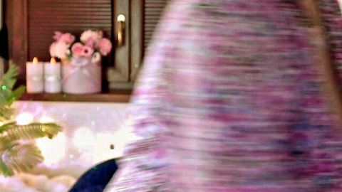 Blurry rear view of woman in colorful knitted shawl in cozy room with pink flowers and candles