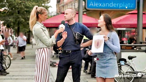 Blonde reporter interviews Asian babe holding giant printed pussy sign with man on Alexanderplatz street