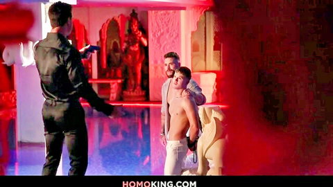 Leather-clad daddy points gun at shirtless young gay hunk in erotic red-lit temple.