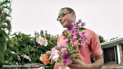 Smiling Black man in pink shirt holding purple and pink flowers outdoors, tropical vibe