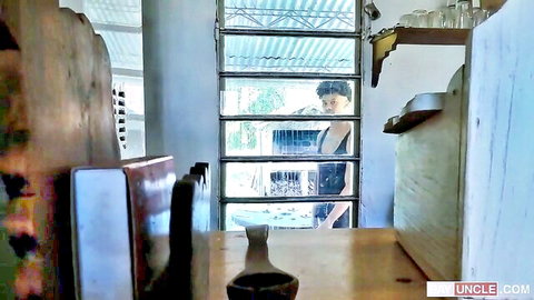Voyeur shot of muscular Asian guy in tank top washing dishes at sink through window