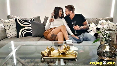 Brunette Latina in mini skirt and sweater flirts intimately with tattooed bearded hunk holding wine glasses on sofa