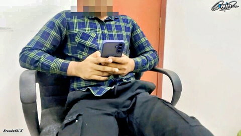 South Asian man in plaid shirt sits legs spread holding phone in office chair