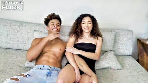 Shirtless muscular black stud with dreads and curvy brunette babe smiling teasingly on couch