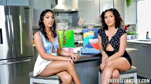 Two sexy Latina brunettes in skimpy crop tops and shorts sit teasingly at kitchen counter
