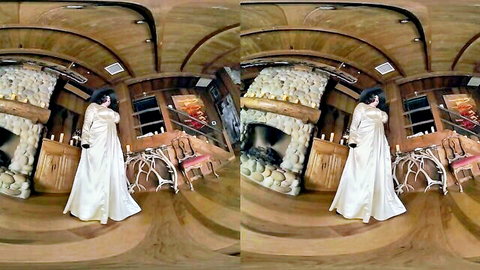 Brunette babe in flowing white gown posing seductively by fireplace in VR cabin