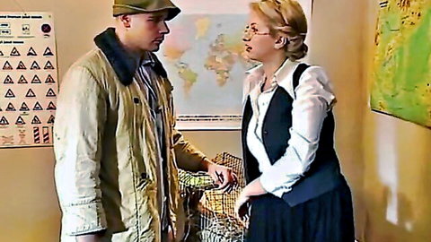 Mature blonde teacher in glasses and uniform chats closely with man in classroom