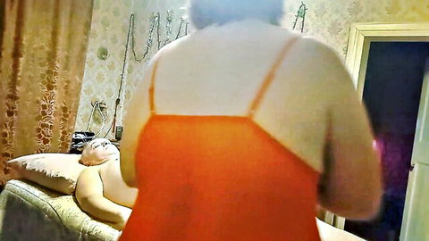 Chubby mature woman in orange dress stands over older man on couch in brothel scene