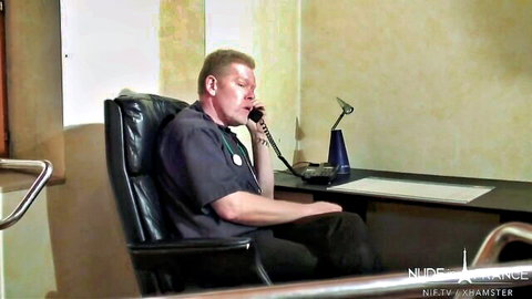 Balding doctor in scrubs sits in leather chair talking on blue phone in office