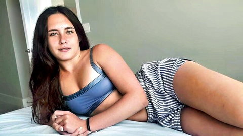 Slim Latina woman in blue sports bra and striped shorts lounging seductively on bed