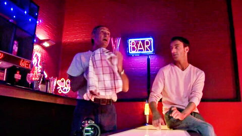 Two clothed men chatting behind neon-lit bar counter with red walls and drinks