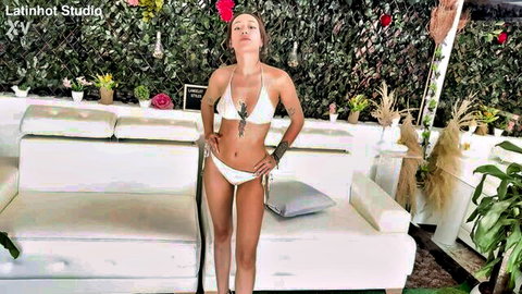 Sexy brunette Latina in tiny white bikini posing hands on hips showing cleavage and curves poolside