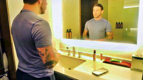 Tattooed muscular man in gray shirt poses in front of bathroom mirror reflection