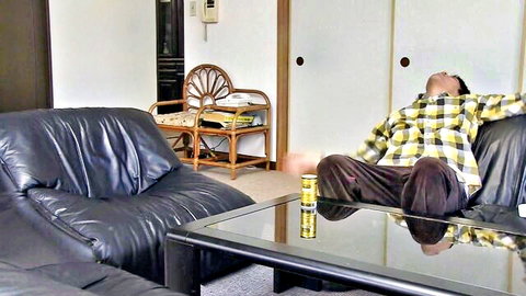 Relaxed Japanese man lounging on black leather sofa in living room with beer can on glass table