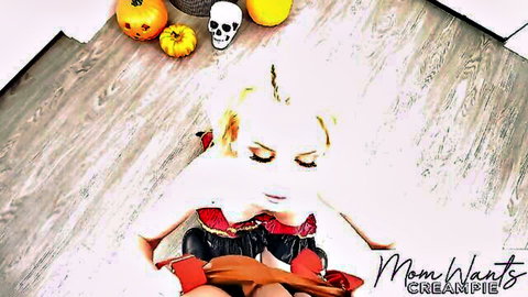 Blonde MILF in sexy red corset Halloween costume teases on floor with pumpkins and skull.