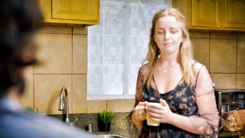 Blonde MILF in sheer floral dress holds mug as man approaches in kitchen