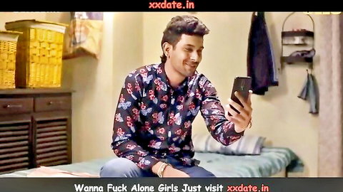 Smiling Indian guy in floral shirt sits on bed holding phone with 'Wanna fuck alone girls just visit xxdate.in' promo text