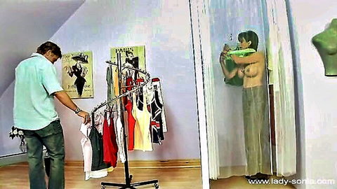 Busty blonde MILF with huge tits topless in changing room curtain as man browses lingerie