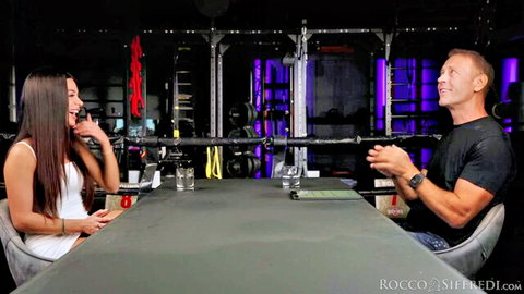 Petite Asian teen with small tits interviews Rocco Siffredi smiling at gym table