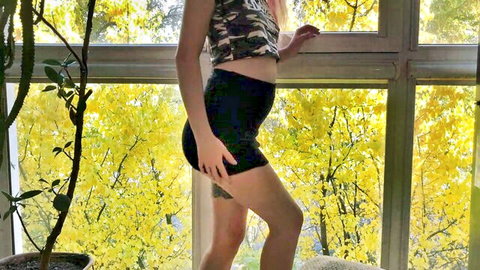 Blonde babe in camo tank top and tight black shorts poses by window
