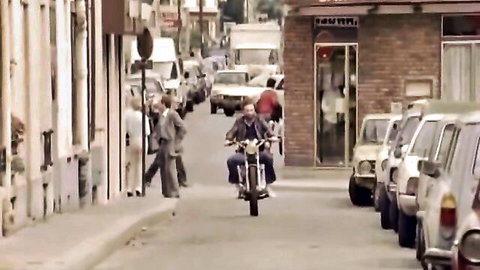 Vintage suited man riding motorcycle down narrow urban street with parked classic cars