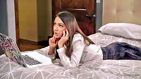 Surprised sexy teen in plaid skirt and open blouse lies on bed talking on phone with laptop