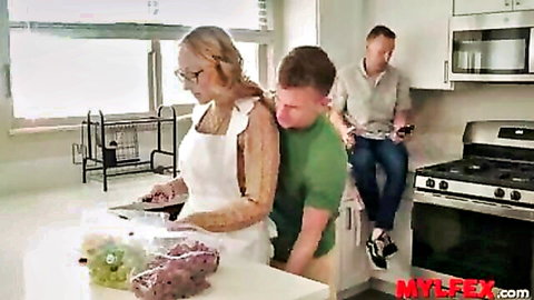 Blonde MILF housewife in glasses and apron hugged tightly from behind by young stud in kitchen as husband watches