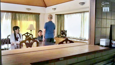 Bald Asian man stands behind table with seated woman in white blouse and young man in living room