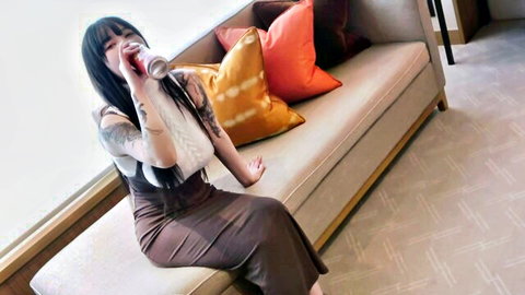 Tattooed Asian woman in sexy dress sipping drink on couch