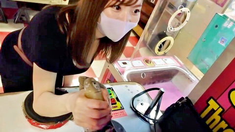 Masked Japanese woman in black dress bends over claw machine, seductive JAV pose