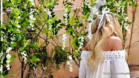 Blonde Kayla in off-shoulder white lace dress under floral arch