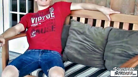 Young muscular guy in red Virtue shirt and ripped jean shorts lounging on porch swing teasing bulge