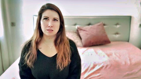 Brunette woman with cleavage in black off-shoulder sweater sitting on pink bed