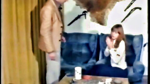 Brunette woman in white blouse pleads with suited man standing by blue couch in retro room