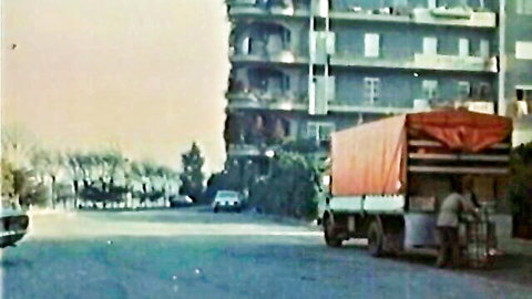 Vintage street scene with man unloading crates from red tarpaulin truck near apartments and cars