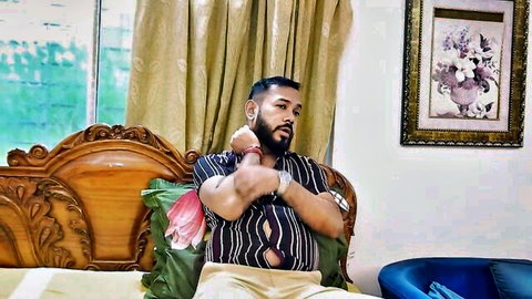 Bearded Indian stud flexing muscular arms on wooden bed in striped shirt and pants