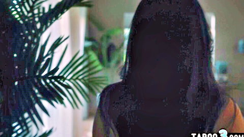 Shadowed long-haired brunette in orange top teases from behind amid plants in taboo scene