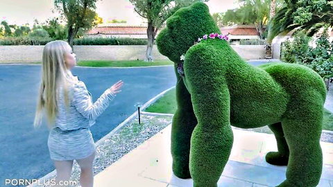 Blonde woman in gray tracksuit posing hand out to giant green moss gorilla topiary outdoors