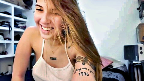 Lolykush smiling in white tank top, blonde hair and arm tattoos on webcam
