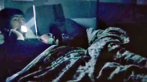 Young Asian man with flashlight peeking at woman under covers in dark bedroom