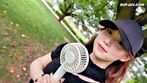Smiling Asian woman in black cap and top holds fan in park
