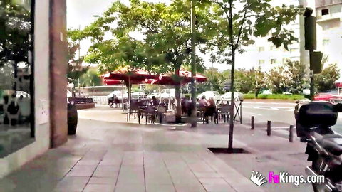 Outdoor street cafe scene with people sitting at tables under red umbrellas on sunny sidewalk