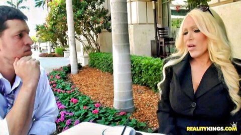 Busty blonde MILF with huge tits in open blazer chats seductively with man outdoors