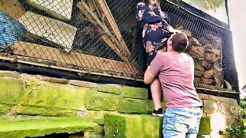 Chubby girlfriend in floral dress lifted over fence in park by boyfriend