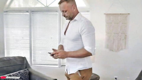 Muscular man in open white shirt checks phone standing by window in living room