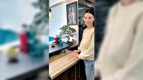 Brunette woman in cream sweater and jeans smiling at phone in modern kitchen