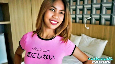 Petite Thai girl in tight pink 'I don't care' shirt smiling teasingly in bedroom