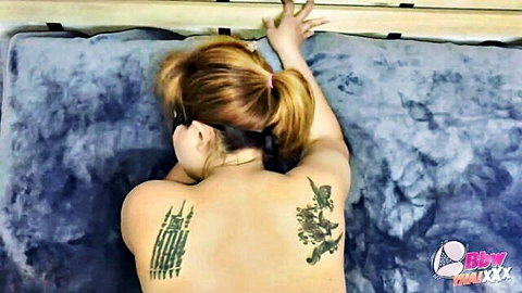 Chubby Thai woman with intricate back tattoos lying nude on bed